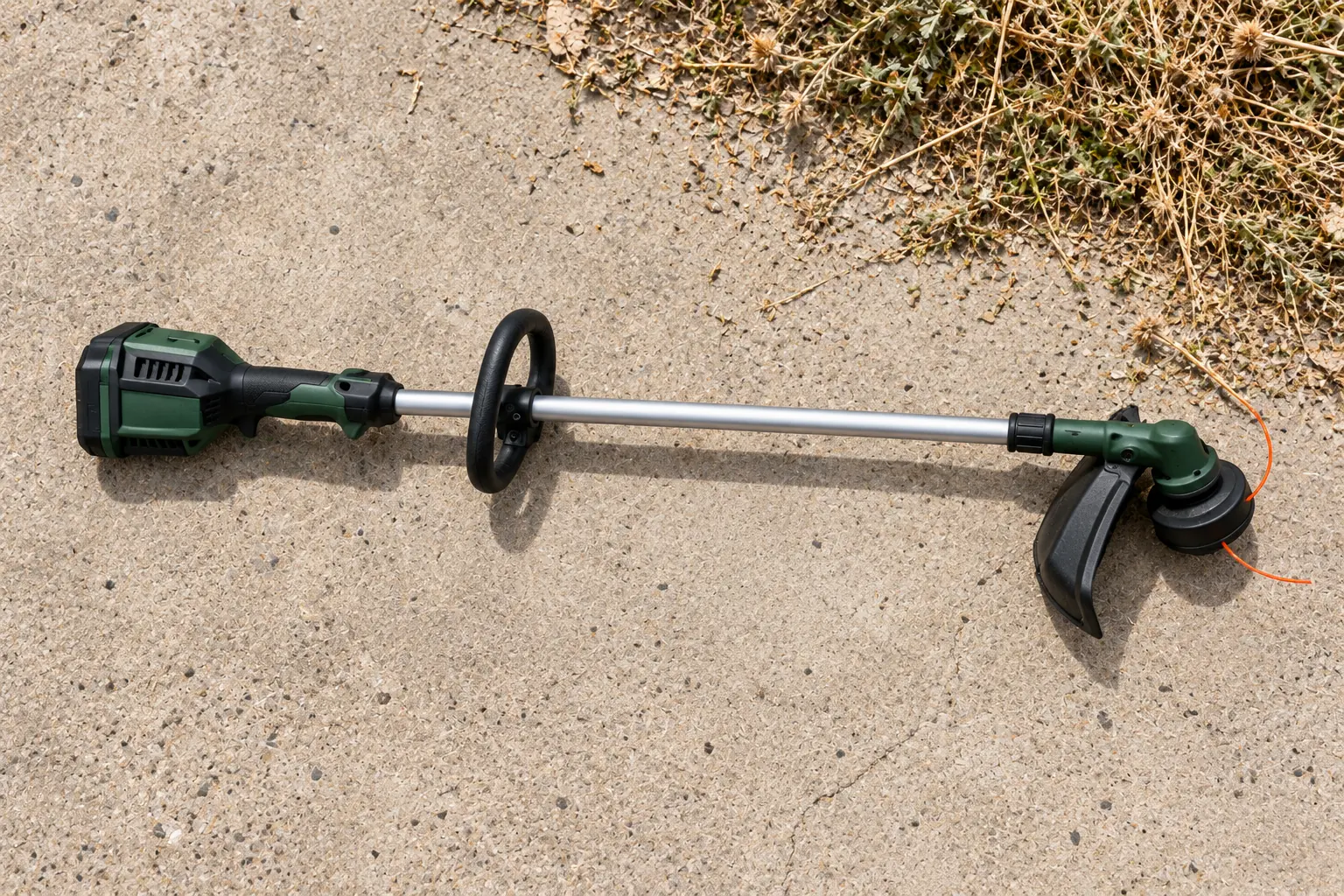 Clean cordless string trimmer resting on concrete beside cut weeds